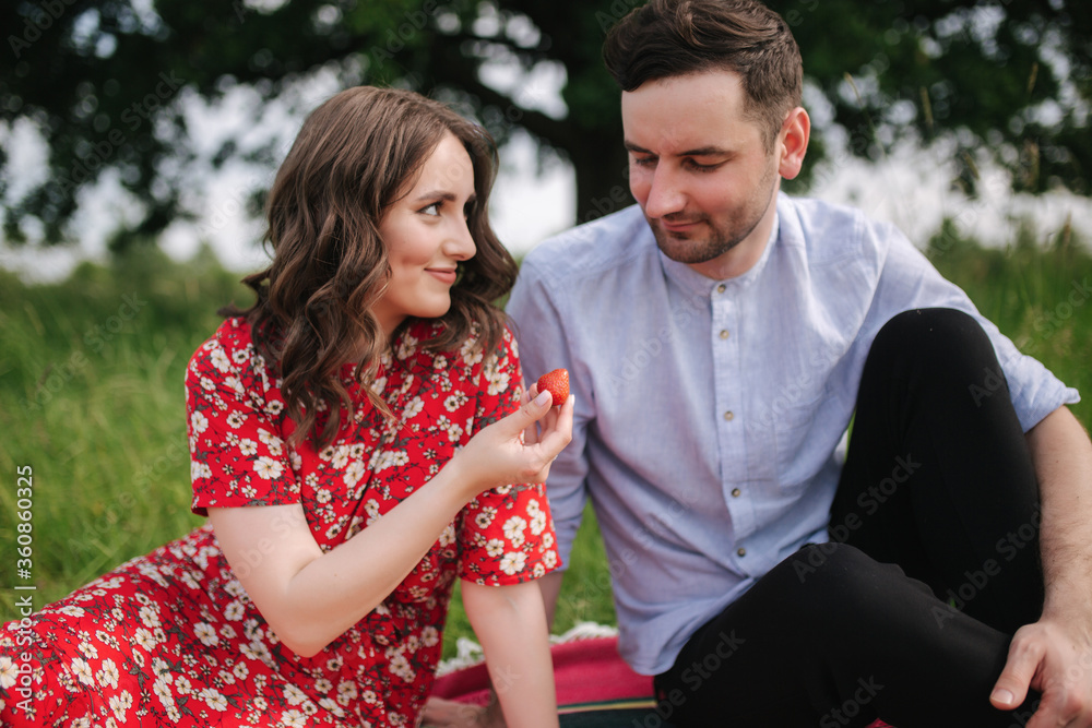 Attactive young woman with handsome man sits outdoors on red blanket. Woman give to man strawberry. Man taste berry Attactive young woman with handsome man sits outdoors on red blanket. Woman give to man strawberry. Man taste berry
