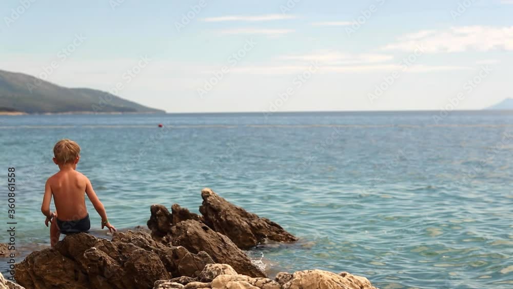 Little boy putting rubber flip-flop under himself while sitting on rock near sea Little boy putting rubber flip-flop under himself while sitting on rock near sea