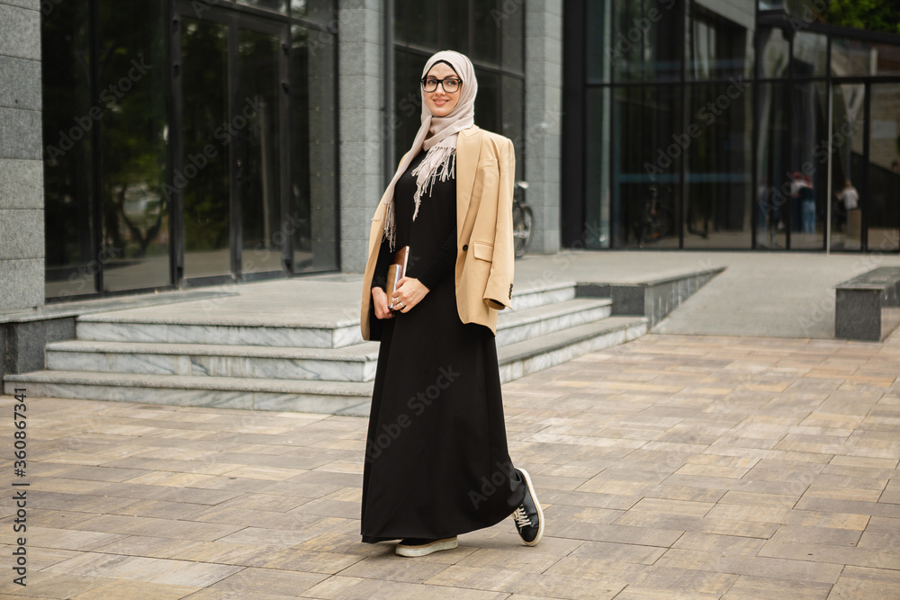 modern stylish muslim woman in hijab business style jacket and black abaya walking in city street with laptop freelancer online remote work business woman in glasses Stock Adobe Stock
