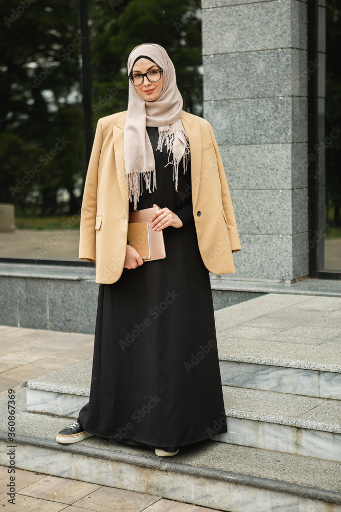 modern stylish muslim woman in hijab, business style jacket and black abaya  walking in city street with laptop, freelancer online remote work, business  woman in glasses Stock-Foto | Adobe Stock