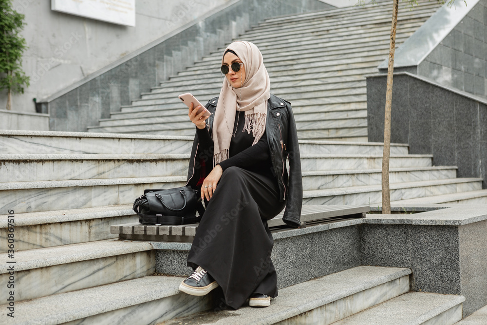 modern stylish muslim woman in hijab, leather jacket and black abaya  walking in city street using smartphone, wearing sunglasses and bag,  fashion style trend Stock Photo | Adobe Stock