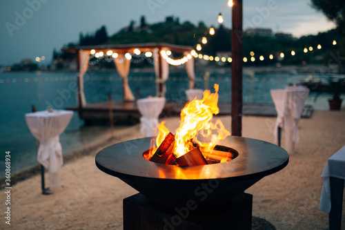 Grill with flames inside. Round table-cooking surface. On the beach, in the background of the gazebo by the water with garlands, in the twilight light.