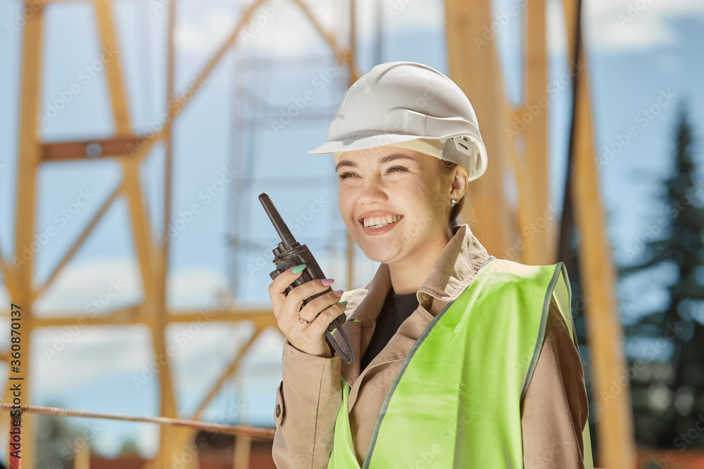 Young girl civil engineer in the background of building construction communicates on walkie with a smile on her face Young girl civil engineer in the background of building construction communicates on walkie with a smile on her face