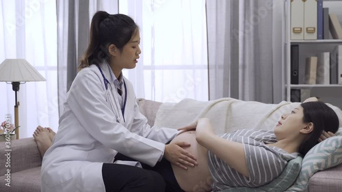 asian korean midwife exanimating bare belly of lying pregnant woman manually with both hands in living room on couch. lady medical staff visit motherhood with naked tummy at home. regular check up. photo