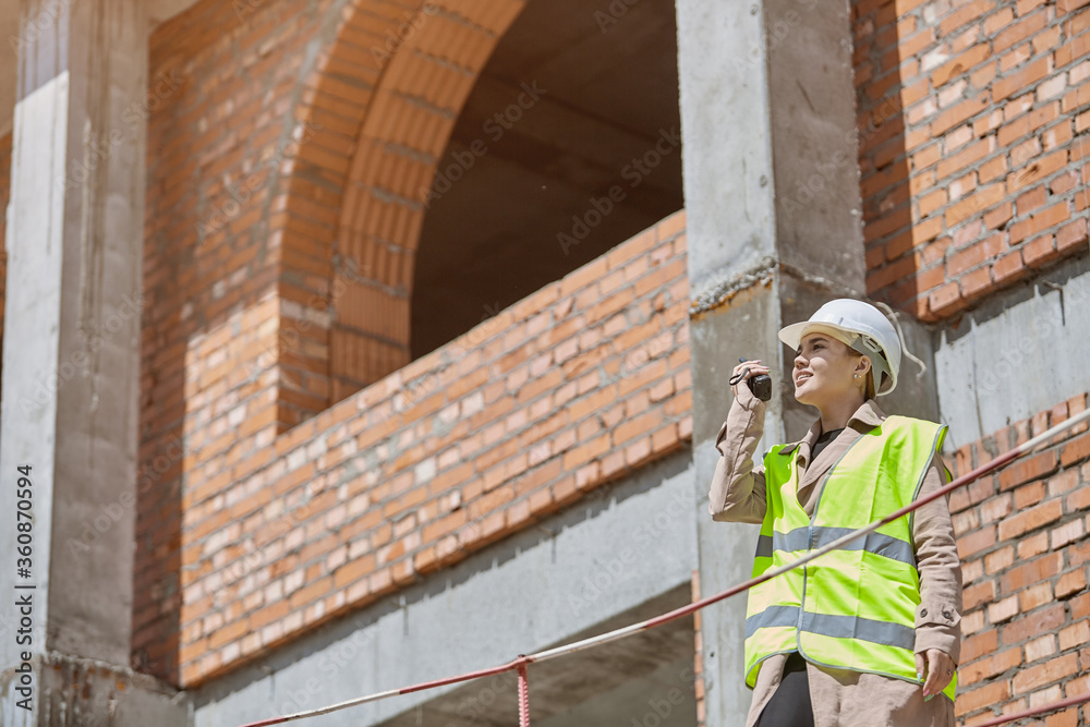 A girl engineer with a walkie talkie in hand on a construction site in a protective vest and hard hat gives instructions