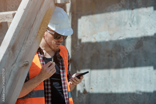 Construction site worker in a protective vest and hard hat, Smoking with a mobile phone in his hands