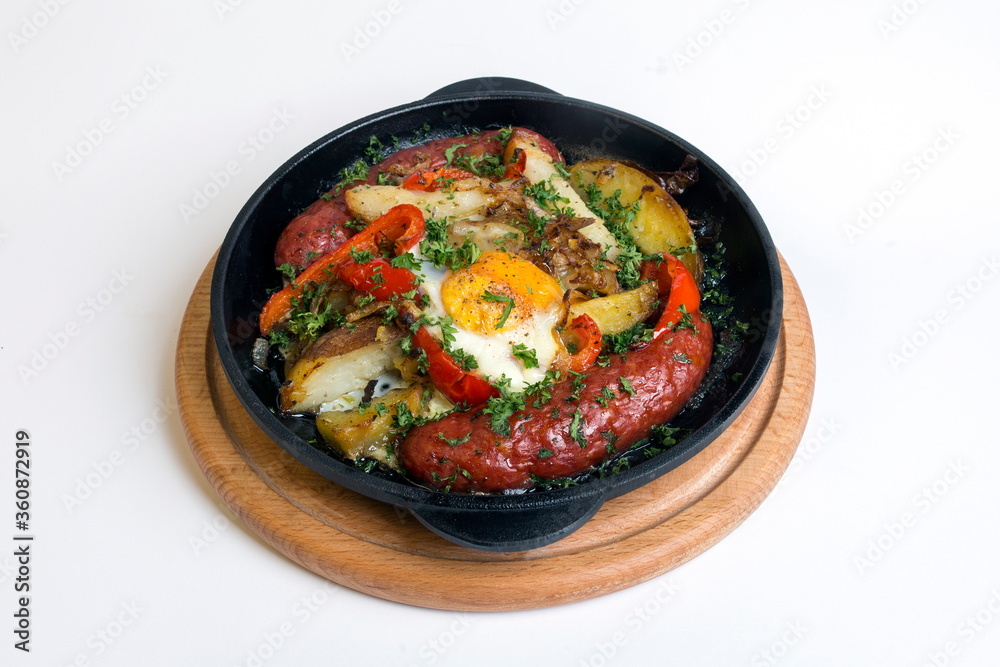 Dish on a black pan, which stands on a wooden round stand. Baked potatoes with sausages and red pepper. Sprinkled with green parsley.