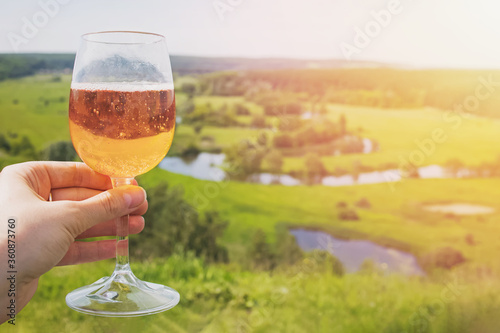 Hand holding a glaa of sider or other alcoholic sparkling drink with the view to the beautiful green meadow in countryside photo