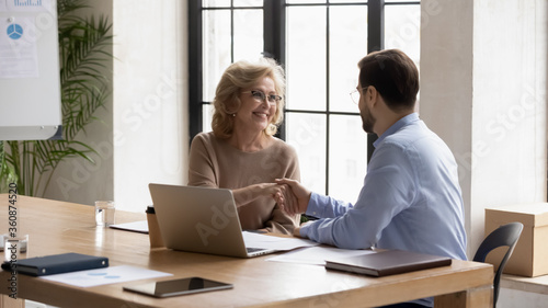 Businessman shaking mature businesswoman partner or customer hand at meeting, making successful deal after negotiations, sitting at table in boardroom, hr manager greeting candidate on job interview