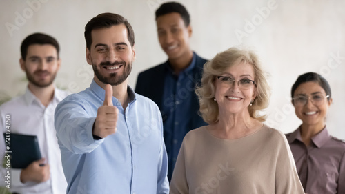 Head shot portrait two team leaders with diverse employees team, smiling middle aged businesswoman and confident businessman showing thumb up, recommending service, good choice, successful career