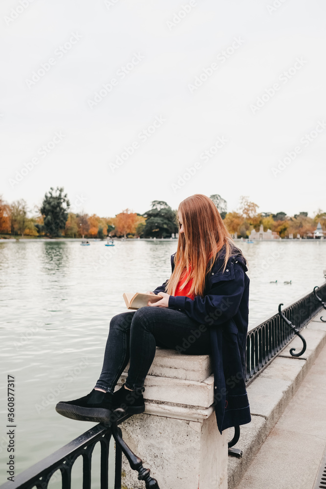 atractiva mujer joven pelirroja con pecas sentada en un banco leyendo y en parque pensativa delante de un estanque