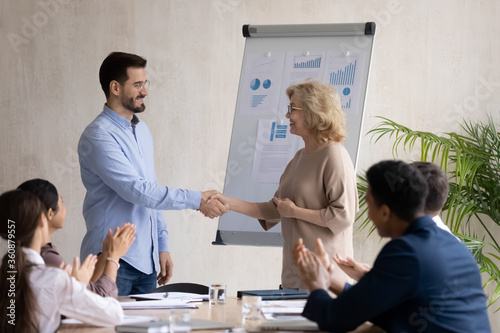 Middle aged businesswoman leader wearing glasses shaking successful employee hand, thanking for good work results, congratulating with job promotion at corporate meeting, hiring new worker