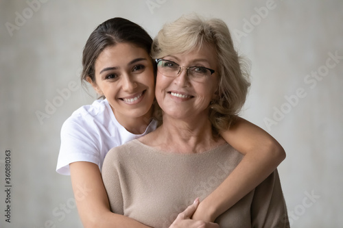 Head shot portrait loving grown up daughter hugging middle aged mother from back, looking at camera, happy mature grandmother and granddaughter posing for family photo on grey background together photo
