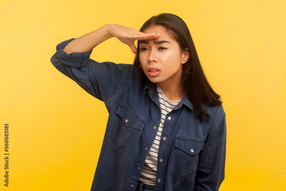 Portrait of curious girl in denim shirt looking carefully far away with hand over eyes, choosing distance, exploring horizon with attentive view. indoor studio shot isolated on yellow background Portrait of curious girl in denim shirt looking carefully far away with hand over eyes, choosing distance, exploring horizon with attentive view. indoor studio shot isolated on yellow background