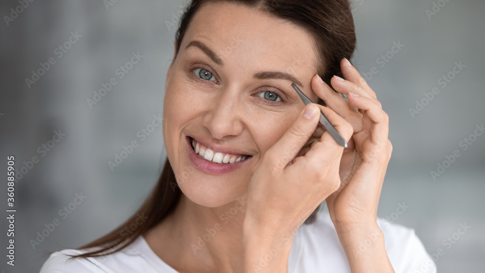 Smiling beautiful young woman look in mirror in home bathroom use tweezers pinch eyebrows, happy millennial female do cosmetics beauty routine procedure, take care of skin, cosmetology concept Smiling beautiful young woman look in mirror in home bathroom use tweezers pinch eyebrows, happy millennial female do cosmetics beauty routine procedure, take care of skin, cosmetology concept