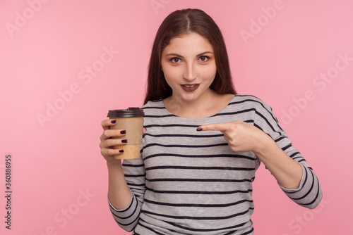Portrait of attractive brunette woman in striped sweatshirt pointing at coffee and smiling to camera, drink with caffeine, energy boost in morning. indoor studio shot isolated on pink background