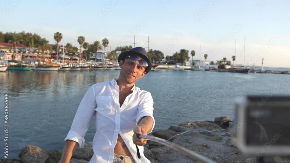 Young man wearing white shirt and hat posing for selfie with handstick while on vacation in exotic place on Mediterranean Sea. Man showing Young man wearing white shirt and hat posing for selfie with handstick while on vacation in exotic place on Mediterranean Sea. Man showing