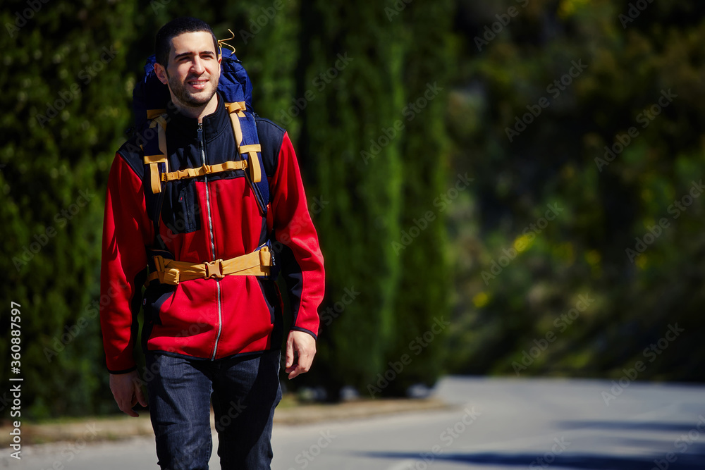 Half length portrait of handsome man tourist with backpack on the back walking on the paved road in the woods during his vacation holidays, young male in sports jacket in sport hike activity outdoors Half length portrait of handsome man tourist with backpack on the back walking on the paved road in the woods during his vacation holidays, young male in sports jacket in sport hike activity outdoors