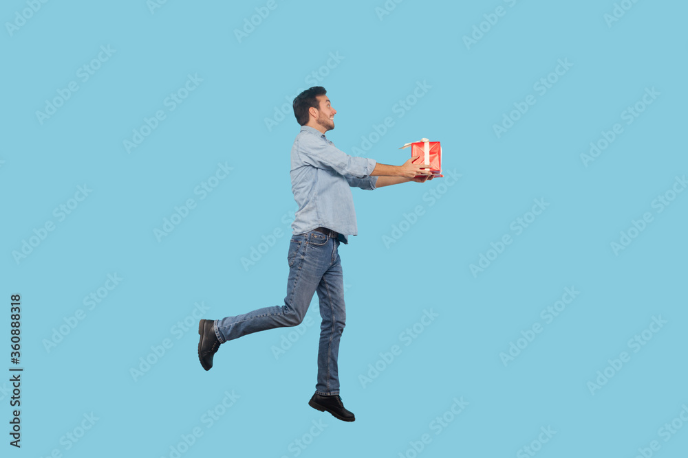 Full length, man in denim outfit flying in air and giving gift, congratulating on birthday, offering holiday surprise, holiday bonus wrapped in box. indoor studio shot isolated on blue background Full length, man in denim outfit flying in air and giving gift, congratulating on birthday, offering holiday surprise, holiday bonus wrapped in box. indoor studio shot isolated on blue background