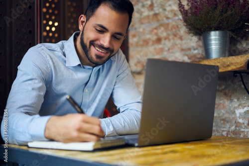Smiling attractive male economist making financial report counting amount of sales using information from corporative database working hard in coworking space using modern laptop computer and wifi photo