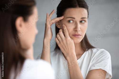 Anxious young woman look in the mirror worried about wrinkle or acne on unhealthy skin, upset unhappy millennial female examine squeeze pimple on face, cosmetology, skincare concept photo