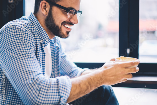 Smiling bearded man enjoying starting day with aroma coffee for breakfast in cozy cafe interior, cheerful hipster guy in casual wear recreating during leisure time sitting in cafeteria holding mug