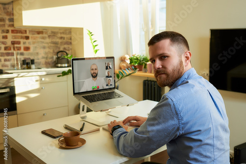 Back view of a male employee who distracted during a talk to his colleagues about a business by a video conference on a laptop computer at home. A multiethnic business team at an online meeting. © Roman Tyukin