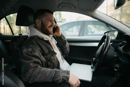 A happy man with a beard doing business calls on his smartphone inside a car, a laptop lies on his lap. A guy smiling stopped his car to immediately remotely solve tasks at work in social distance. © Roman Tyukin