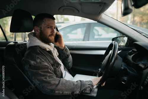 A man with a beard in casual clothes doing business calls on his smartphone inside a car, a laptop lies on his lap. A guy stopped his car to immediately remotely solve tasks at work in social distance