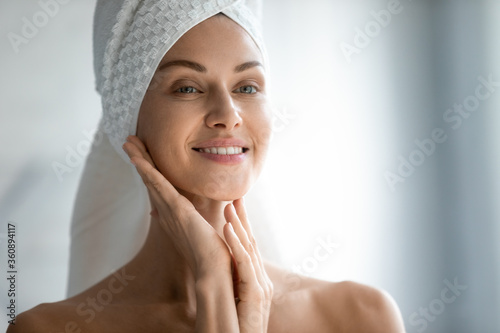 Beautiful young woman after shower look in mirror massage touch soft healthy glowing face skin, happy millennial female apply beauty products do daily facial procedures or treatment, skincare concept