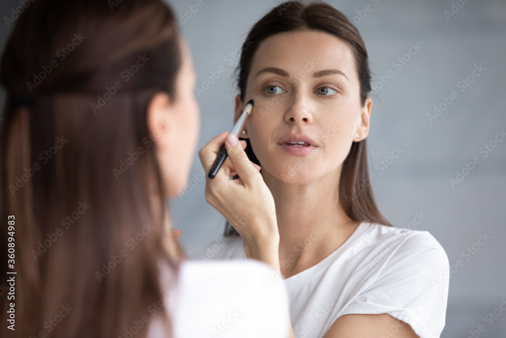 Pretty young brunette woman look in mirror use makeup brush get ready in bathroom in the morning, beautiful millennial female prepare do make up at home bath, cosmetics, beauty routine concept