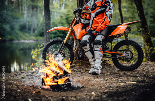 Charming young female racer wearing motocross outfit on her bike and warming up next to a bonfire in the woods. Cropped image
