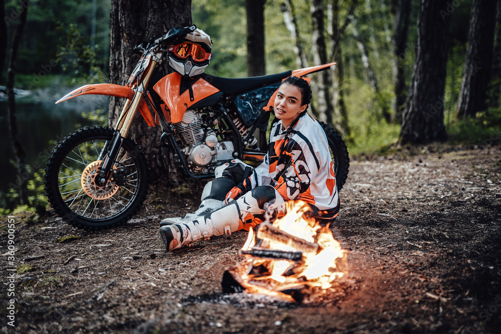 Naklejka premium Beautiful young female racer wearing motocross outfit, warming up sitting next to the bonfire