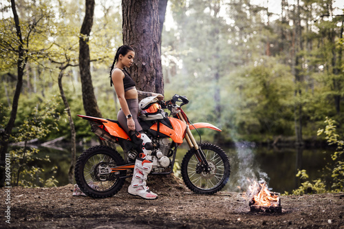 Charming young female racer wearing motocross outfit with semi naked torso sitting on her bike next to the bonfire in the woods