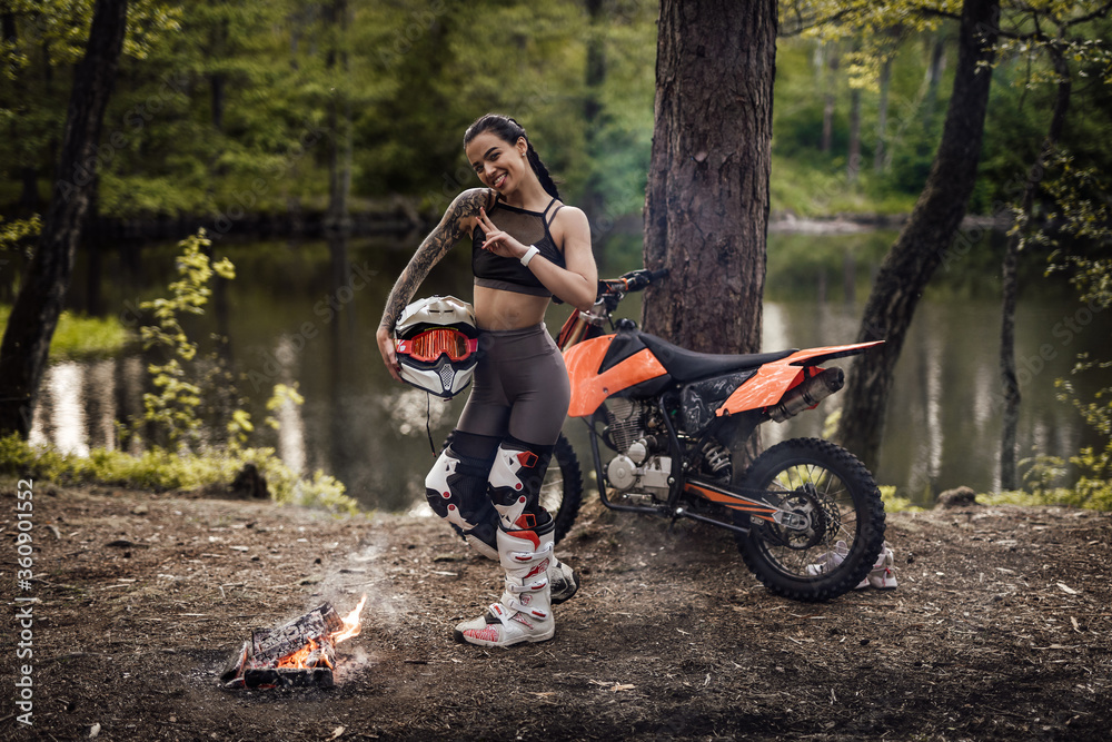 Cheeky racer girl wearing motocross outfit holding helmet and posing on a camera next to the bonfire in the woods