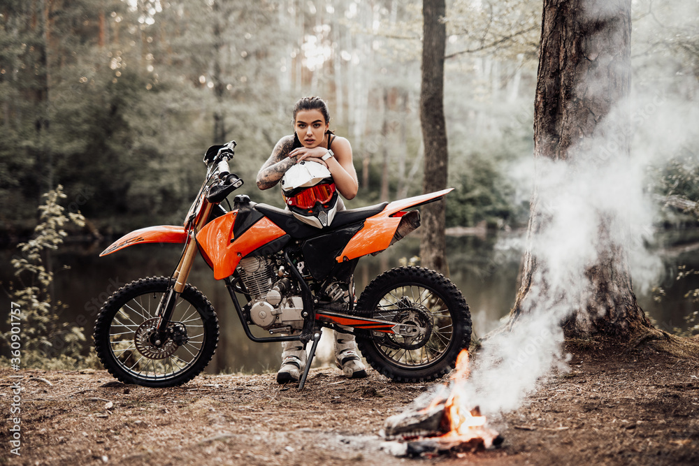 Sensual tattooed racer girl wearing motocross outfit with semi naked torso leaning on her bike and looking on camera in the woods Sensual tattooed racer girl wearing motocross outfit with semi naked torso leaning on her bike and looking on camera in the woods