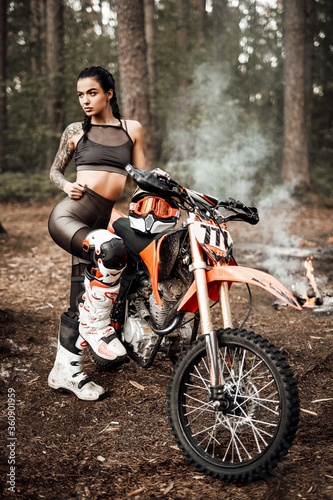 Fototapeta Naklejka Na Ścianę i Meble -  Sensual tattooed racer girl wearing motocross outfit with semi naked torso posing next to her bikein the woods
