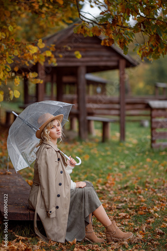 a blonde with blue eyes in a hat and a beige cloak sits on a wooden park bench under an umbrella, smiles, enjoys nature, it is raining. The colors of autumn. Autumn mood. Rainy weather. gold autumn
