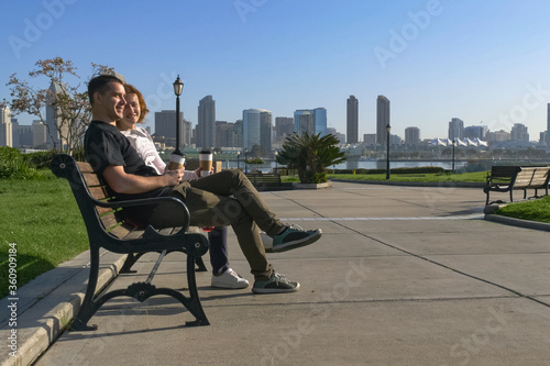 Young couple is sitting on a bench in the city, the sun is shining, they are talking and laughing, watching the distance and dreaming. In the background is the downtown San Diego with skyscrapers.