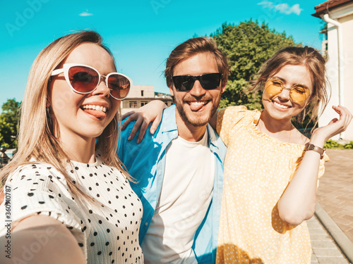 Group of young three stylish friends in the street.Man and two cute girls dressed in casual summer clothes.Smiling models having fun in sunglasses.Women and guy making photo selfie on smartphone