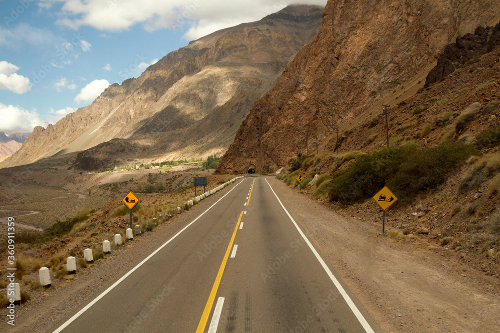 Fototapeta premium Travel. Asphalt highway in the mountains and valley on the road to mountain Aconcagua in Mendoza, Patagonia Argentina. The road crosses a tunnel in the rocky hill.