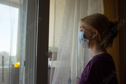 Pretty girl stands by the window. Stylish lady in the mask. Quarantined woman. Coronavirus theme