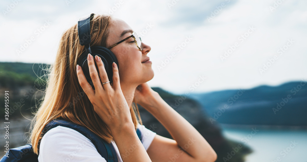 tourist with long hair and hipster glasses listens to her favorite music with headphones smiles and closes her eyes enjoying freedom of traveling through natural mountain landscape on vacation