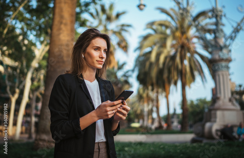 elegant girl in suit holds a smartphone in hand and looks into distance while walking in a Park in Barcelona, girl мanager checks chat with colleagues on mobile phone after office work