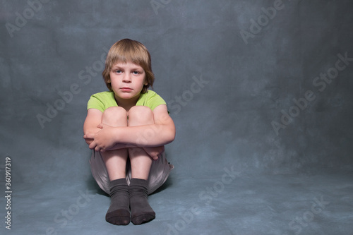 Boy 5 years old, with blond hair, European. The child sits on the floor, hugs his knees, looks at the camera. Gray background, space for copy.