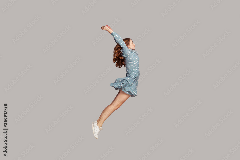 Side view girl in dress levitating hovering in mid-air with raised hands, model looking away concentrated focused, jumping trampoline gaining speed. indoor studio shot isolated on gray background