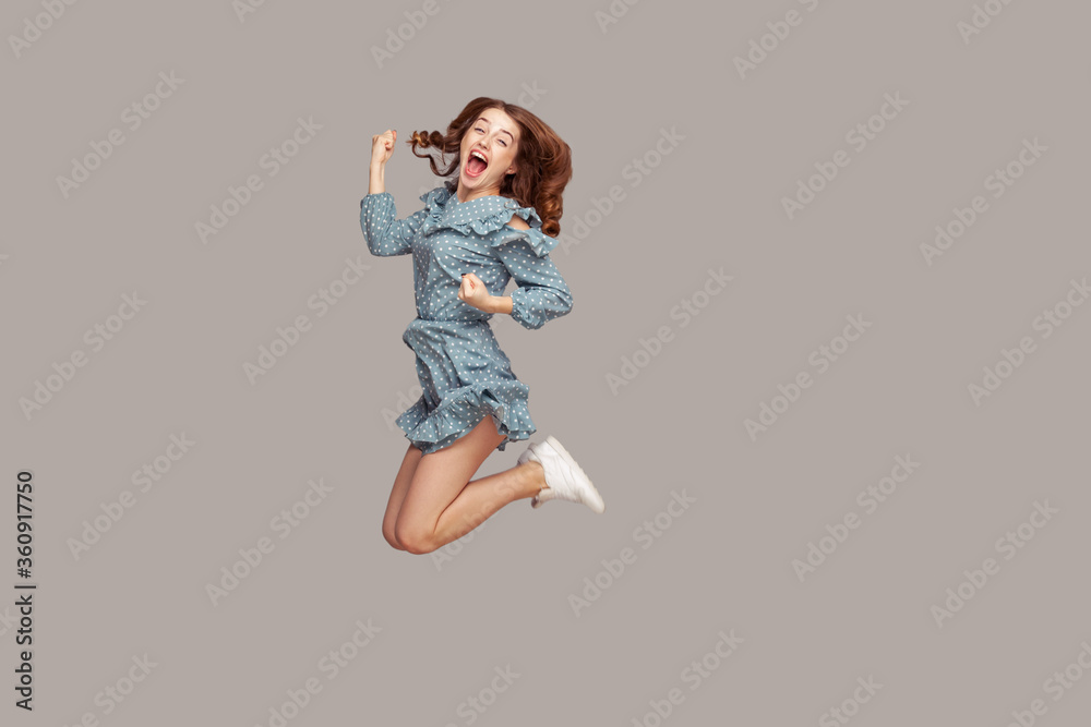 Delighted enthusiastic girl ruffle dress flying mid-air with raised fists shouting for joy, jumping trampoline looking at camera extremely happy, celebrating success. studio shot isolated on gray Delighted enthusiastic girl ruffle dress flying mid-air with raised fists shouting for joy, jumping trampoline looking at camera extremely happy, celebrating success. studio shot isolated on gray