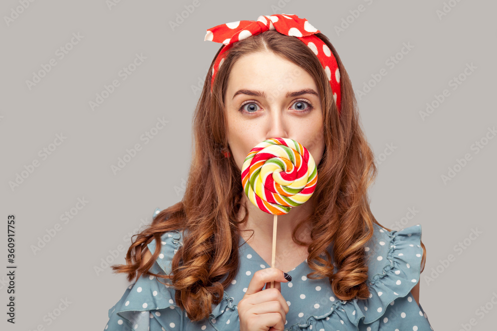 Amazed pinup girl ruffle blouse licking sweet candy looking at camera, eating delicious confectionery lollipop with surprised expression. retro vintage style. studio shot isolated gray background,
