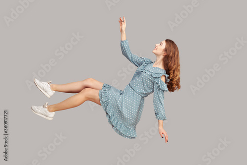 Hovering in air. Cheerful smiling pretty girl in vintage ruffle dress levitating flying in mid-air, looking up happy dreamy and raising hand to catch. indoor studio shot isolated on gray background