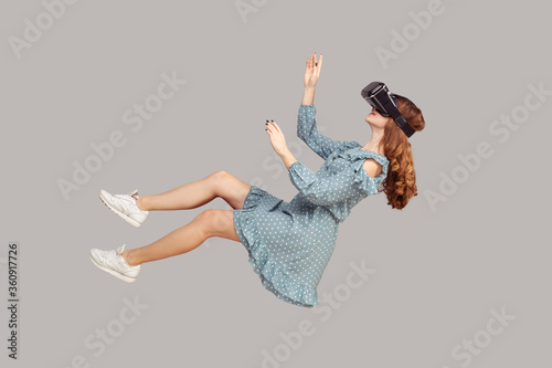 Gamer girl in ruffle dress hovering in air, levitating with virtual reality glasses on head, playing game through vr headset, floating in cyberspace. indoor studio shot isolated on gray background